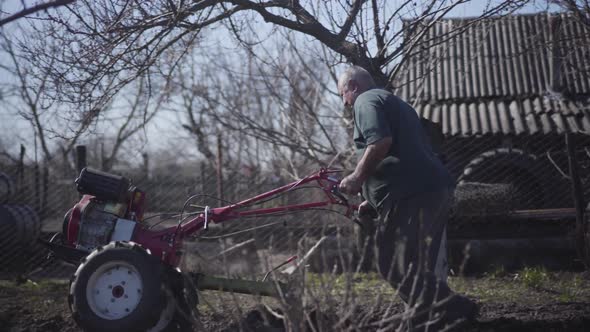 Side View of Mature Caucasian Male Farmer Making Furrows with Furrower. Elderly Man Furrowing Soil