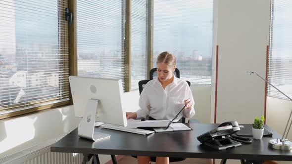 Female Manager Reads Documents and Financial Reports Sitting at the Workplace White Office Interior