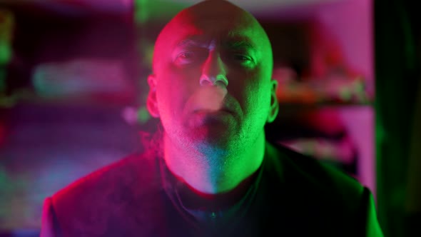 a Bald Man Looks Frowningly at the Camera and Lets Smoke Out of Nose in a Flashing Multicolored Neon