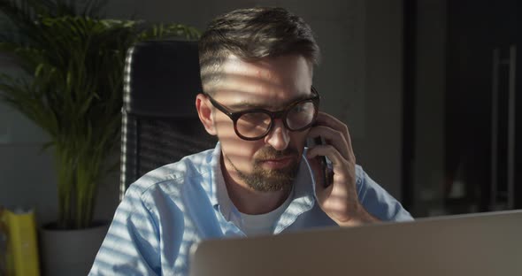 Business Man Using Laptop Talking on Cell Phone Sits at Desk