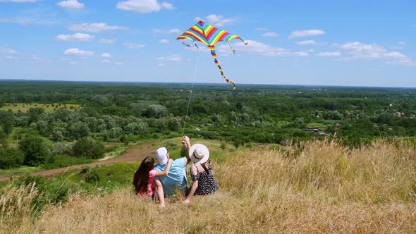 Happy Young Family Sit on Cliff Edge, Fly a Multi-colored Kite, Having Fun, Against Background of