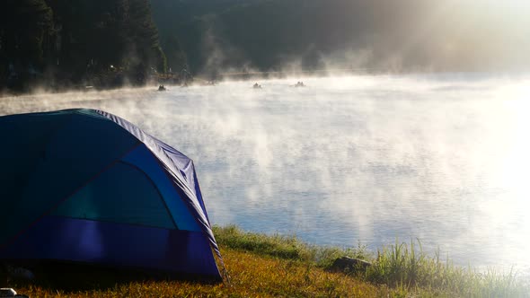 Tent Stands on Shore of Foggy Lake in Early Morning at Dawn in Rays of Sunlight