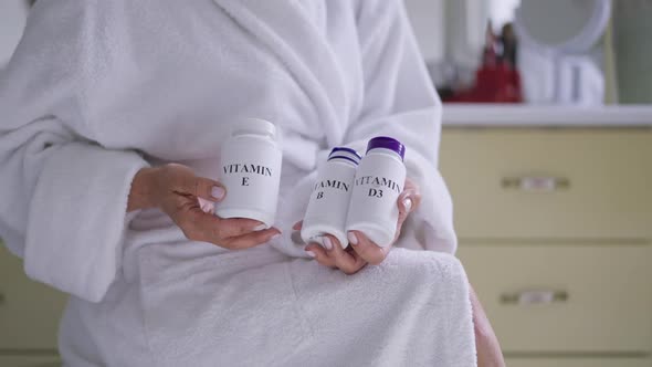 Vitamin Bottles in Hands of Unrecognizable Mature Caucasian Woman in White Bathrobe Sitting in