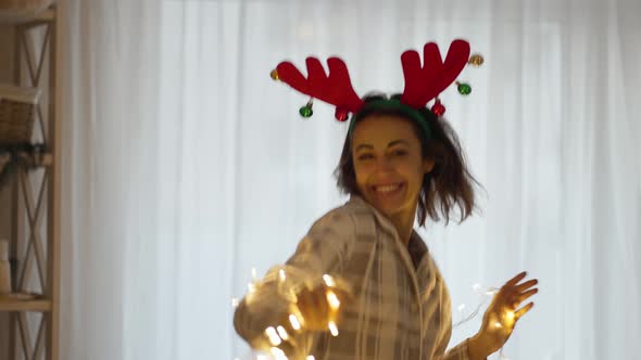 Young Cheerful Pretty Woman Wearing Reindeer Antlers Headband Dancing with Christmas Lights at Cozy
