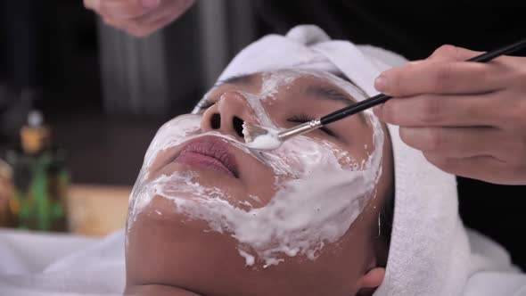 Beautician use cream mask on face of young Asian woman with brush in spa salon.