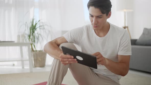 Smiling Young Man Surfing Internet on Tablet While Sitting on Exercise Mat During Relaxing After