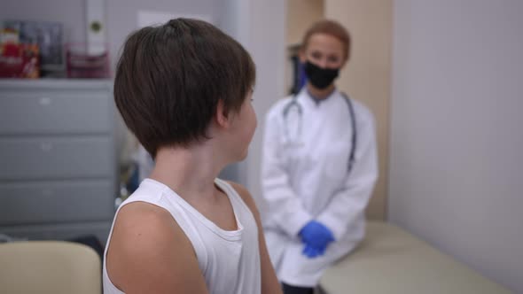Charming Positive Caucasian Boy Looking Back at Blurred Doctor Pointing at Medical Patch on Shoulder
