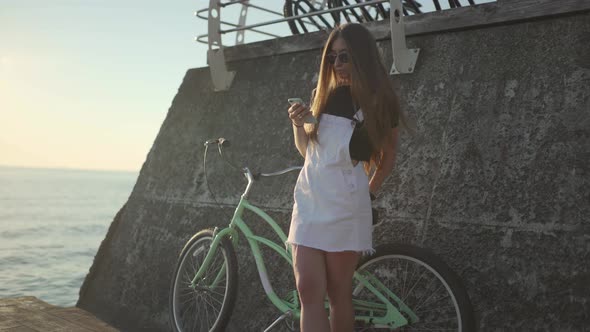 Young Attractive Woman Uses a Smartphone and Riding Vintage Bike Near the Sea During Sunrise or