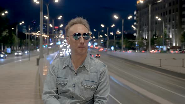Mature Man Poses in Sunglasses at Parapet in Evening City