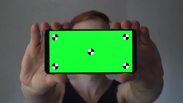 Man holding a smartphone with green screen and tracking points vertically in front of him.