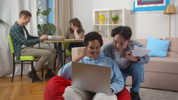 African Woman Sitting on Bean Bag and Watching Funny Video with Korean Friend on Laptop