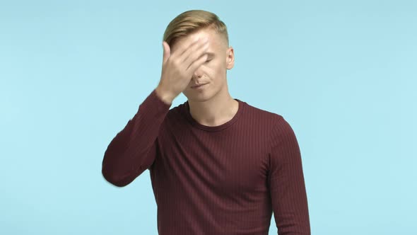 Annoyed and Tired Blond Man Looking Fed Up Making Face Palm and Staring at Camera Standing Over Blue