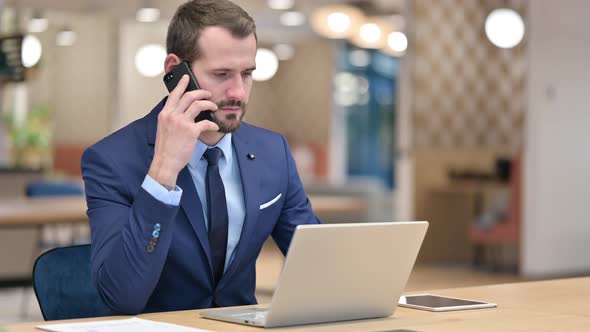 Businessman with Laptop Talking on Smartphone in Office 