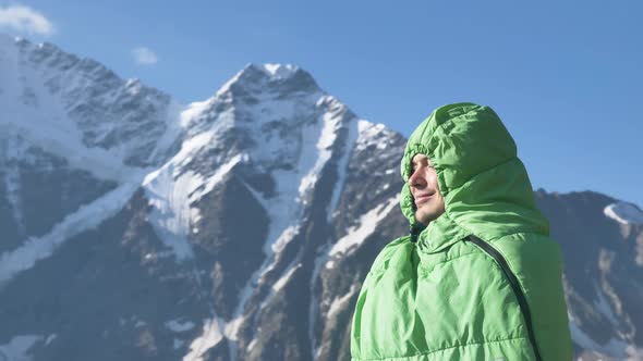 Happy Male Tourist Wrapped in a Sleeping Bag Stands Against the Backdrop of High Snowy Mountains and