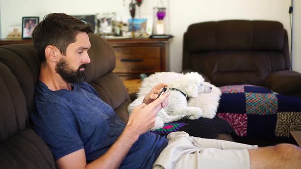 Caucasian man relaxing on sofa using smart phone, dog in background, medium shot