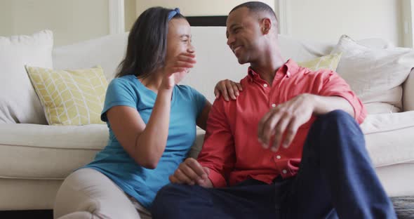 Happy biracial couple sitting on floor in living room, embracing and looking at camera