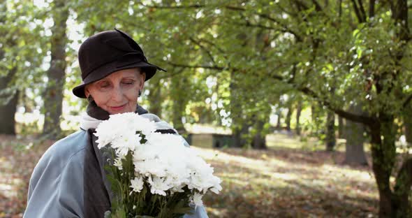 Elegant Elderly Woman in Coat and Hat Sniffing a Bouquet of Flowers with Smile