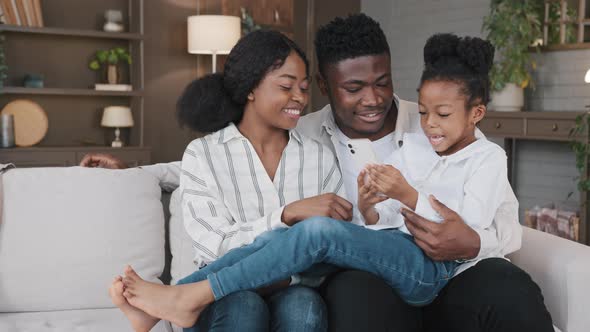 Happy African Family Parents with Cute Schoolgirl Kid Child Relax on Sofa Using Funny Smartphone App