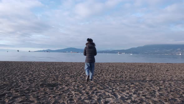 Youg woman wearing black beanie, black puffy coat and jeans on a beach walking towards the sea on a