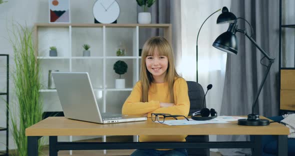 Schoolgirl in Yellow Sweater Sitting at the Table at Home and Looking at Camera