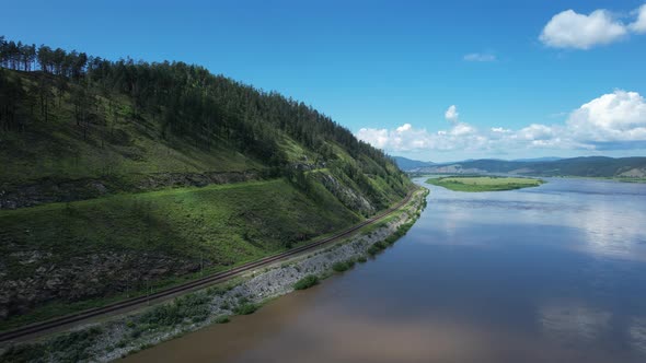 Flying Above a Glacial River in Siberia Russia with Green Mountains and Railway Track in the