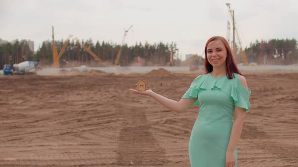 Young Woman with Small Wooden House in Hand Stands on Construction Site