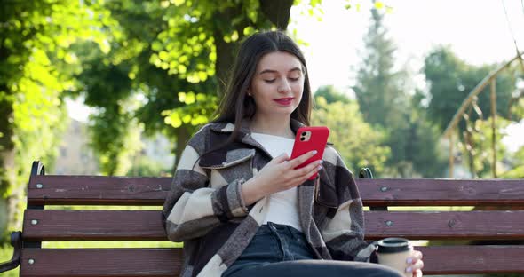 Pretty Young Woman in Trendy Casual Outfit Sits on a Bench in the City Park Actively Texts Messages