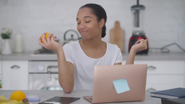 Young Positive Woman Juggling Orange Sitting at Kitchen Table and Typing on Laptop Keyboard