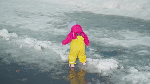 Toddler runs through puddles melting snow in a yellow rubber jumpsuit. Top view