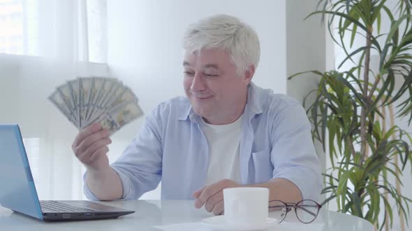 Happy Senior Man Sitting at the Table Holding Pack of Dollars and Smiling at Camera