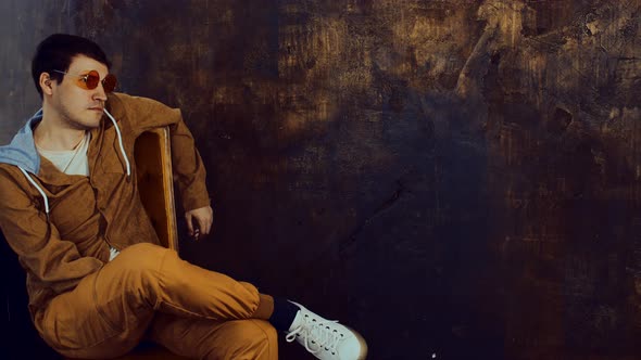 Stylish Man in Bright Glasses Sits Crossing One Leg Over Other on Chair Near Shabby Brown Wall