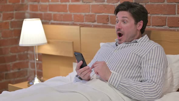 Middle Aged Man Celebrating Success on Smartphone in Bed