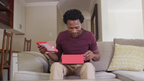 Happy biracial man sitting on sofa, having valentines video call and opening present