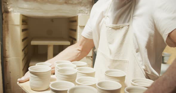 Smiling senior caucasian man wearing apron taking fired pots from kiln at pottery workshop