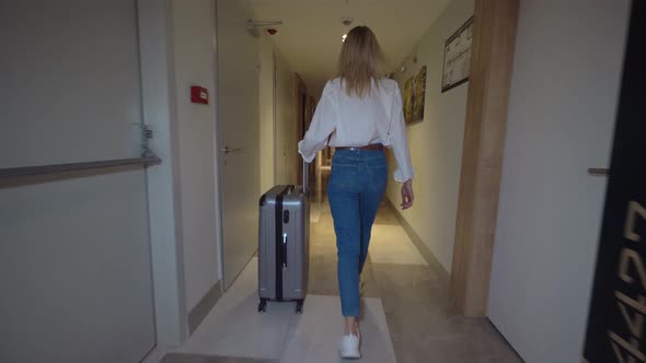 woman walks down corridor of hotel and rolls suitcase with luggage on wheels.