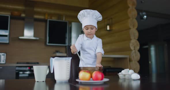 Little Boy in Chef's Costume Makes Apple Pie Dough with Whisk in the Kitchen