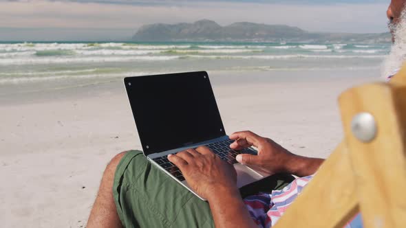 Mixed race senior man sitting on sunbed and using a laptop at the beach