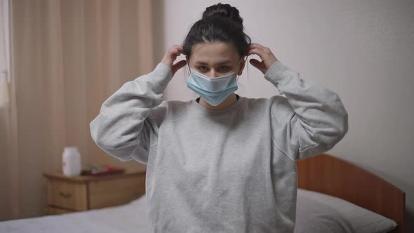 Young Woman Putting on Coronavirus Face Mask Looking at Camera Sitting on Bed Indoors