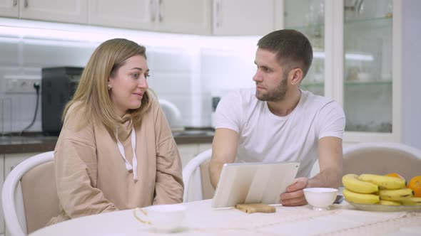 Relaxed Young Caucasian Couple Surfing Internet on Tablet and Talking at Home