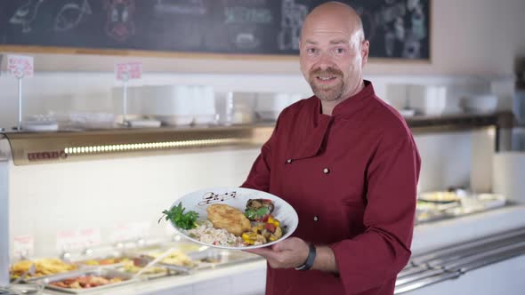 Portrait of Satisfied Bearded Male Cook Showing Plate with Delicious Lunch Looking at Camera Smiling