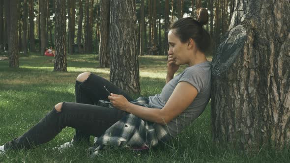 Woman sitting on her skateboard on grass in city park looking at screen on phone 