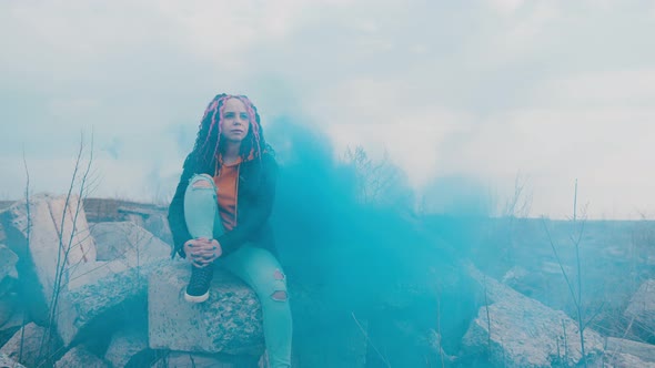 Young Woman Sitting on Destroyed Concrete Blocks with Dissipating Blue Smoke