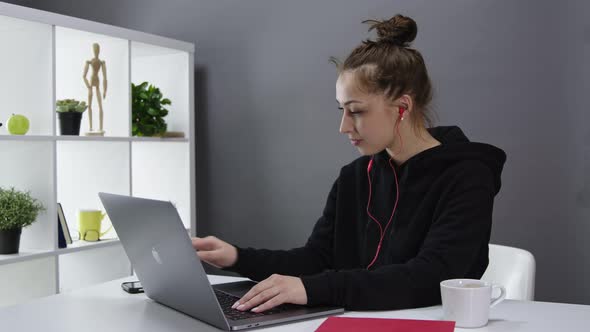 Beautiful Multitasking Girl with Headphones, Hair in Bun Works at Laptop at Home