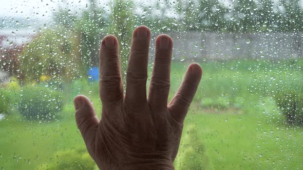 Palm Of Old Man Hand Touches Window Glass In Rain With Drops On Glass Close Up