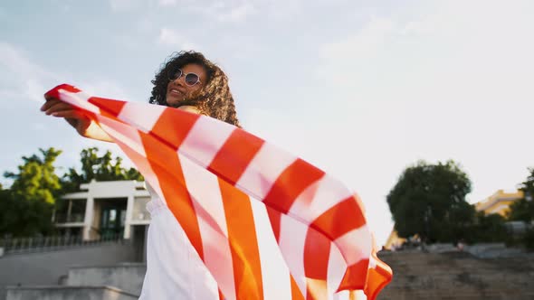 Darkskinned Young Girl in Sunglasses is Smiling and Raising Flag of USA Above Her Head While Posing