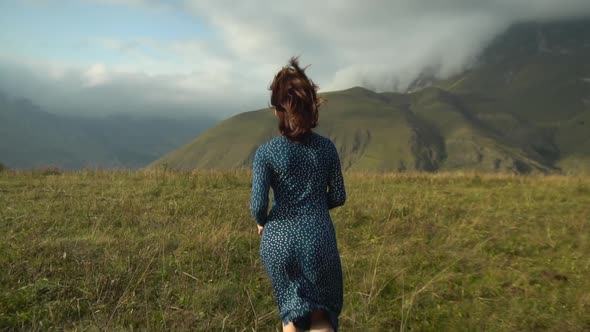 A Young Woman in a Blue Dress Runs Down the Hill Against the Background of the Caucasian Cloudy
