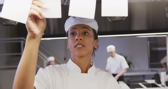 African American female chef working in a restaurant kitchen checking orders, with colleagues workin