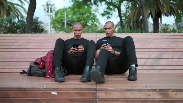 Two confident bald African brothers talking and typing on phone while sitting on the bench