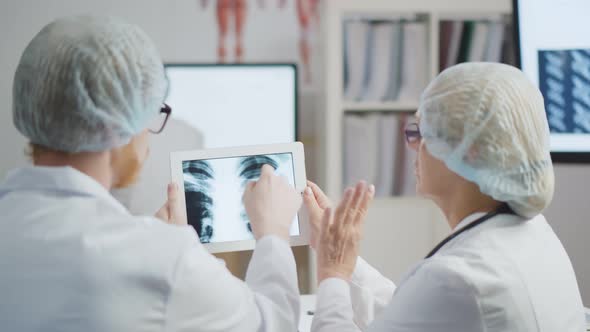 Back View of Two Doctors Having Meeting In Modern Hospital Looking At X Ray On Digital Tablet