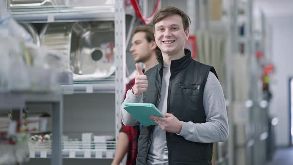 Positive Smiling Worker Looking at Camera Showing Thumb Up As Customer Passing at Background in